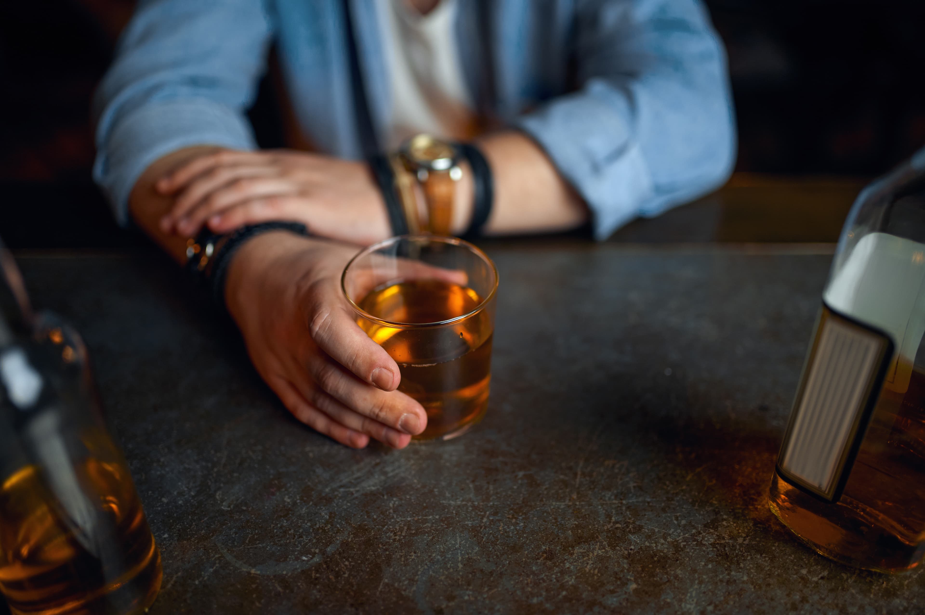 A man's hands grip a glass of whiskey on a dark bar counter with bottles nearby.