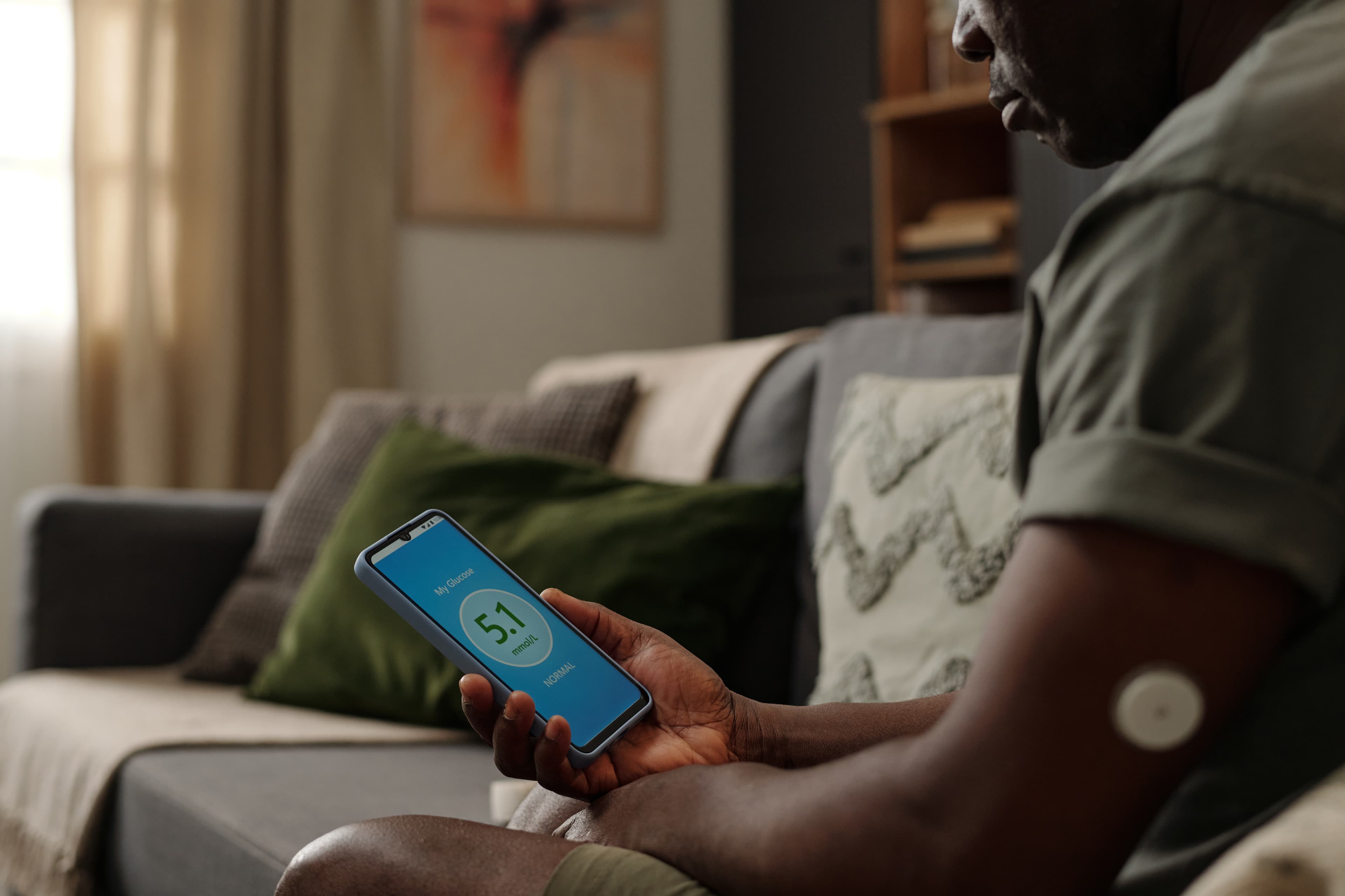 Man sitting on a couch checking a CGM glucose reading on his smartphone while wearing an arm sensor.