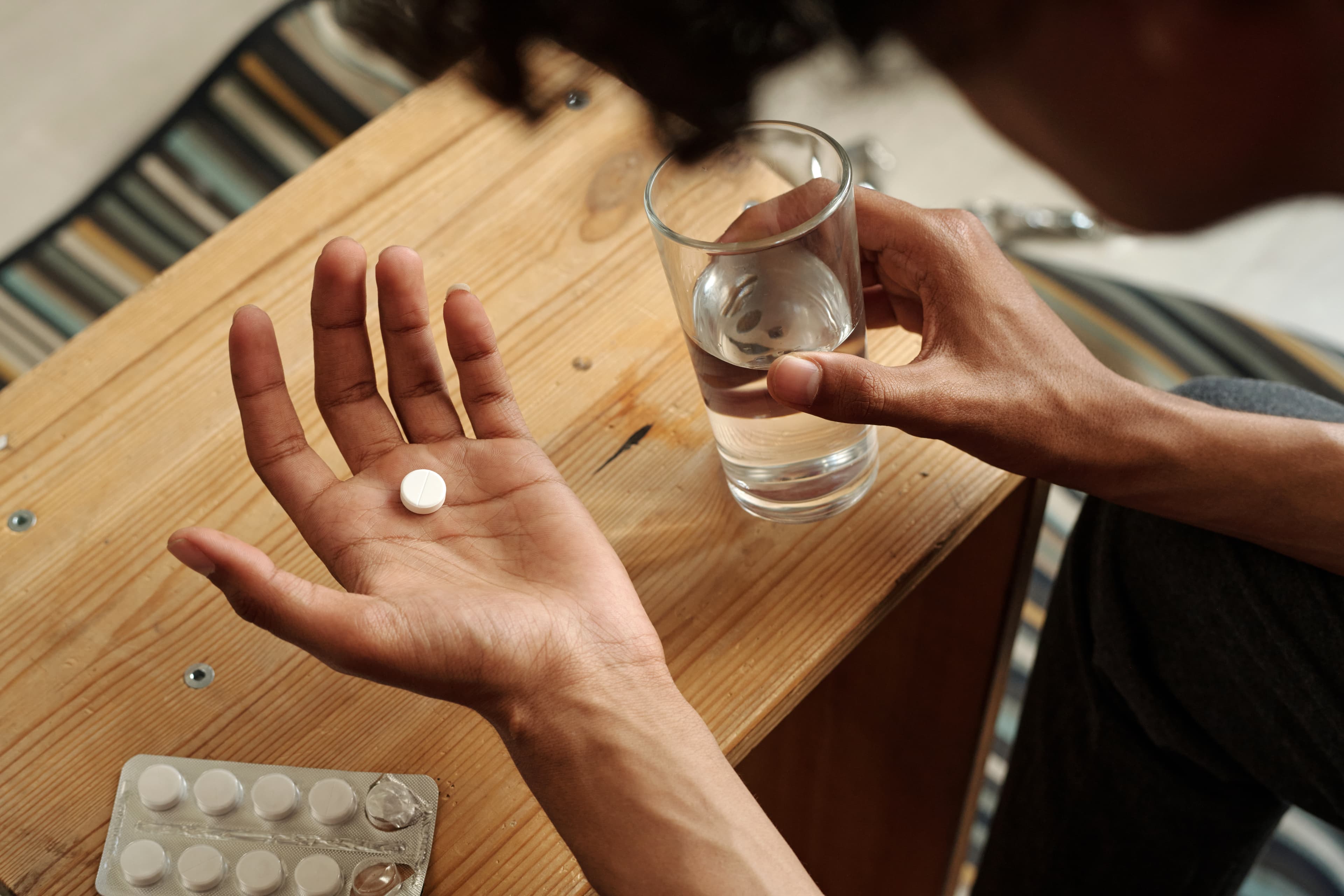 Person holding a single white pill in an open palm alongside a glass of water at a wooden table.