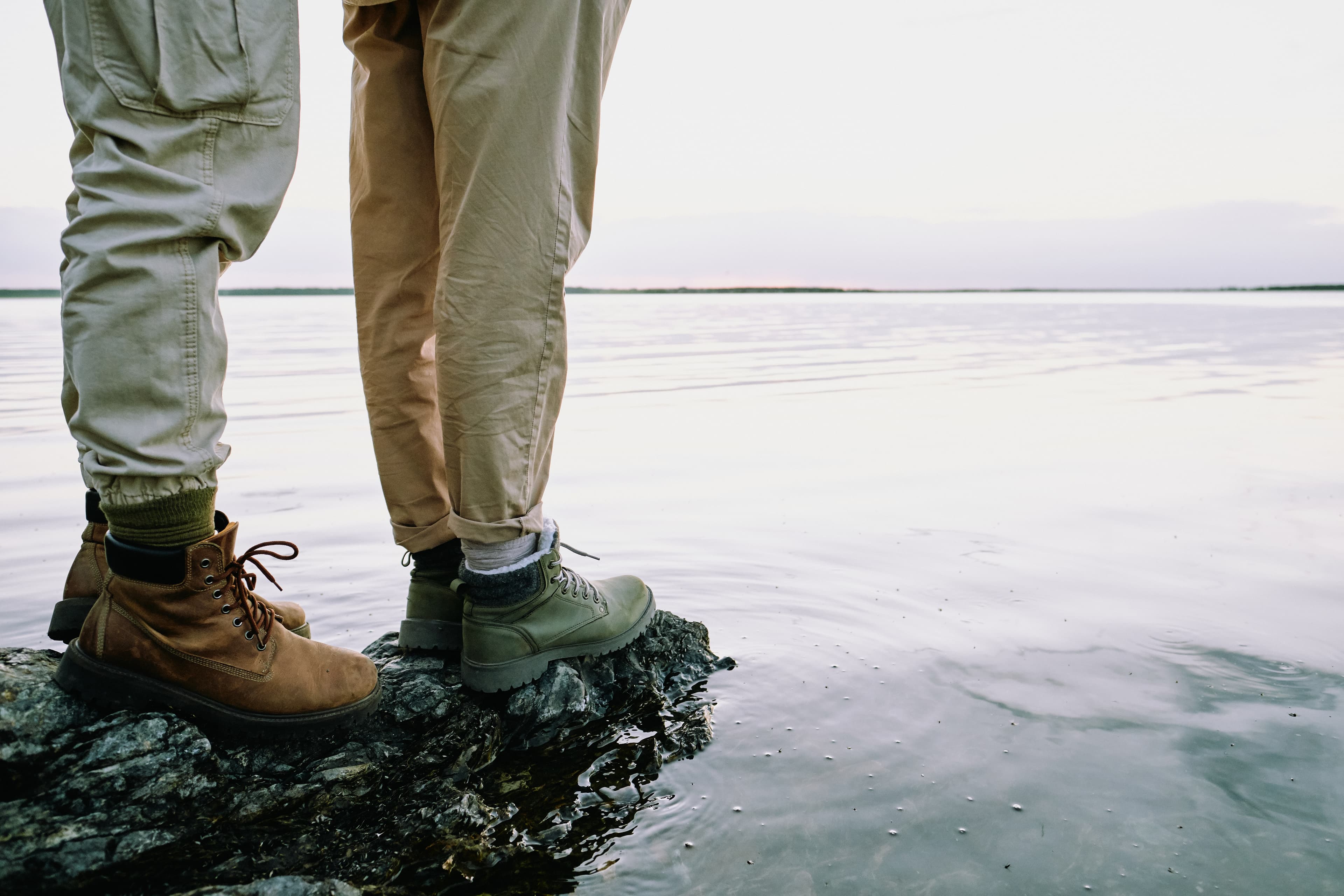 Two people standing on rocks at the edge of a calm body of water, shown from the knees down.