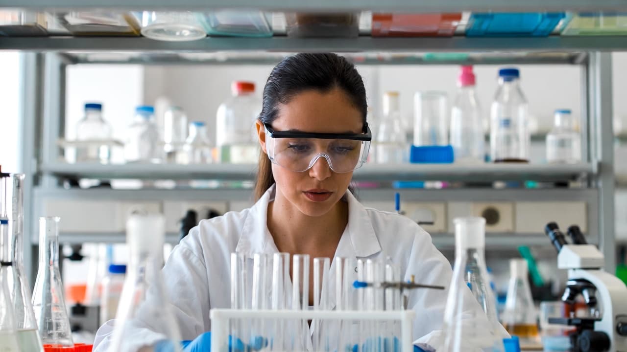 Female scientist in safety goggles examining test tubes inside a laboratory.