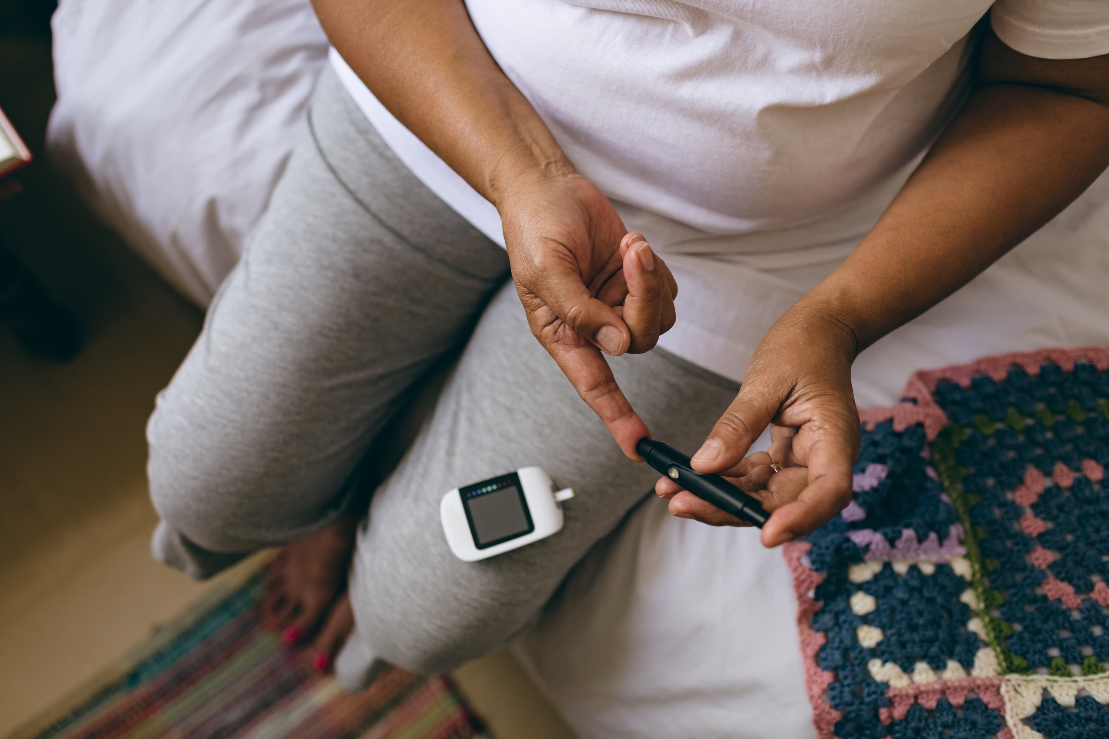 Woman sitting on a bed using a lancet to check her blood sugar with a glucose meter on her lap.