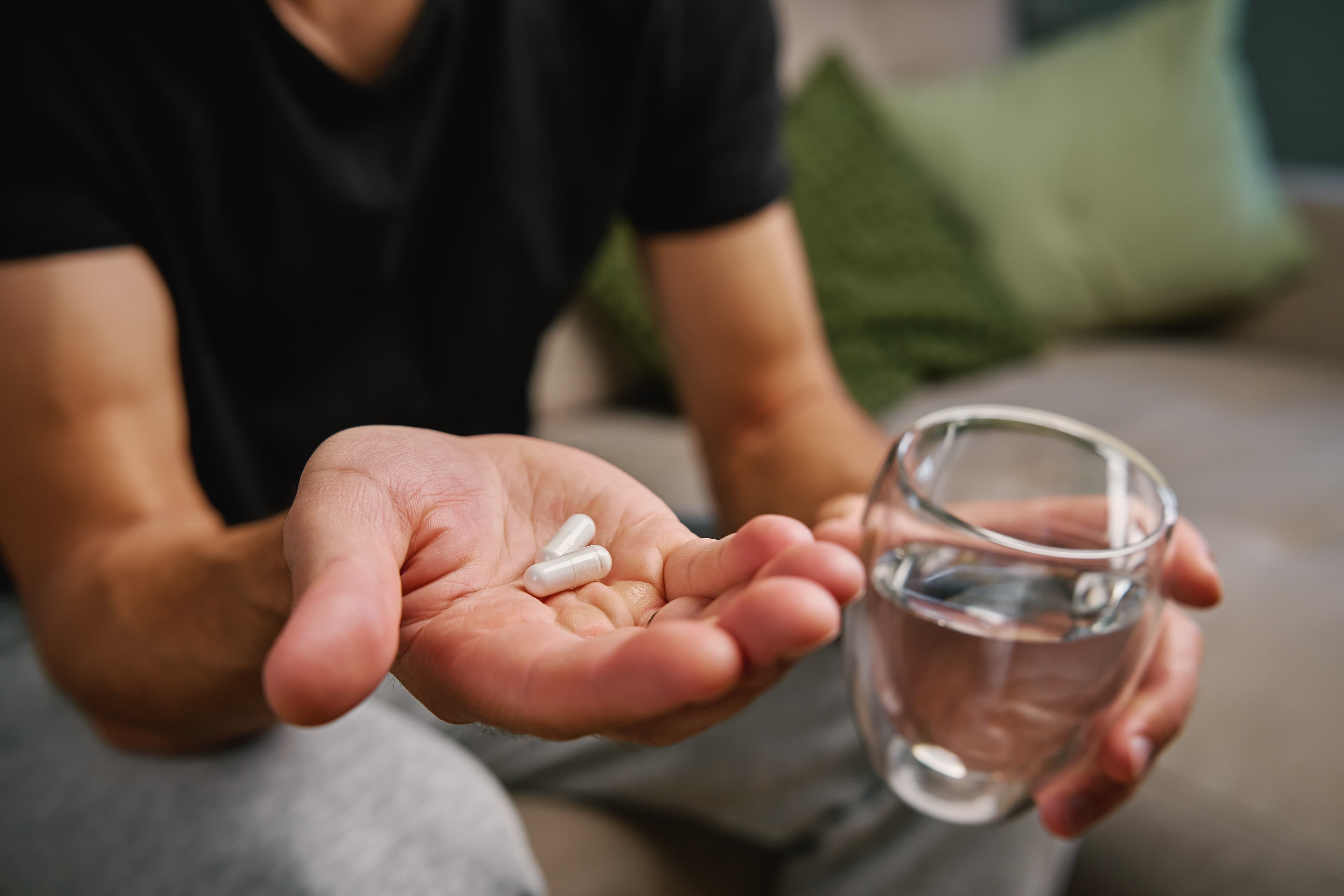 Man's open palm holding two white capsules beside a glass of water, seated on a couch.