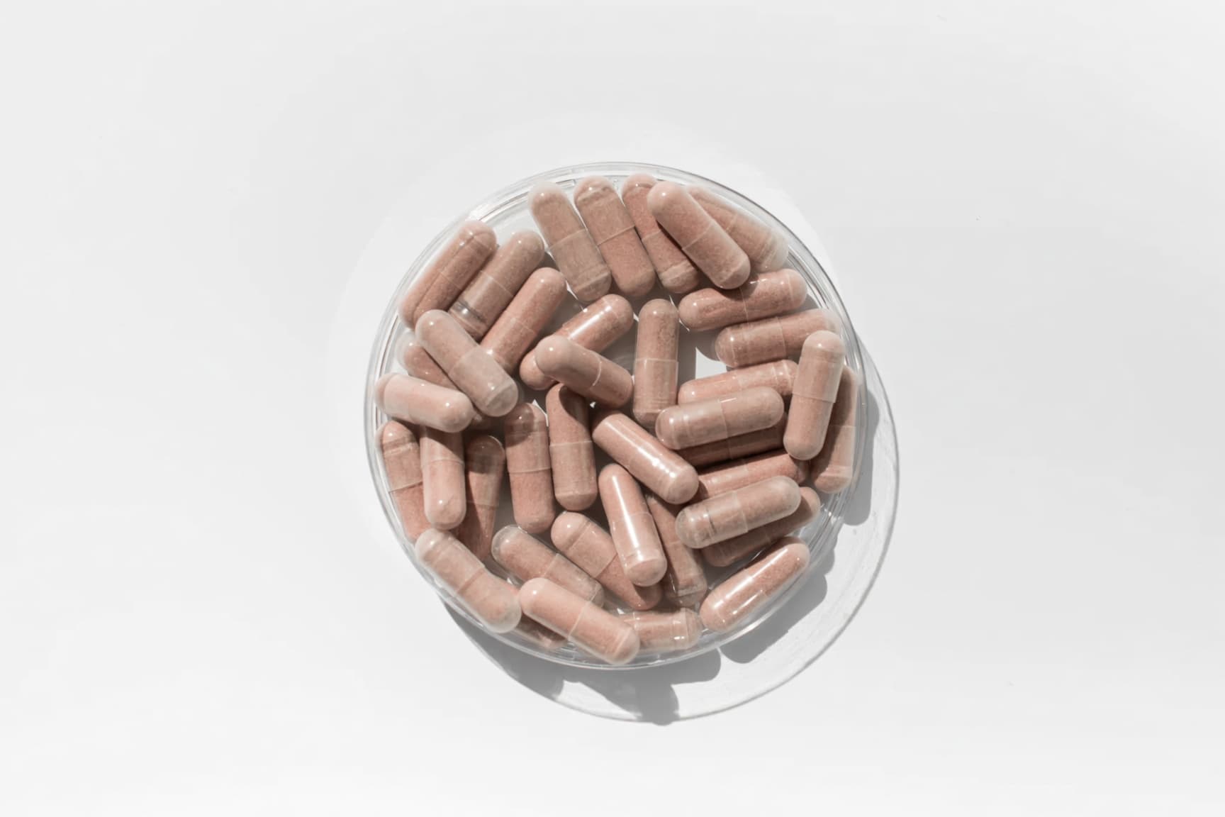 An overhead view of pink-brown supplement capsules piled in a clear glass bowl on a white surface.
