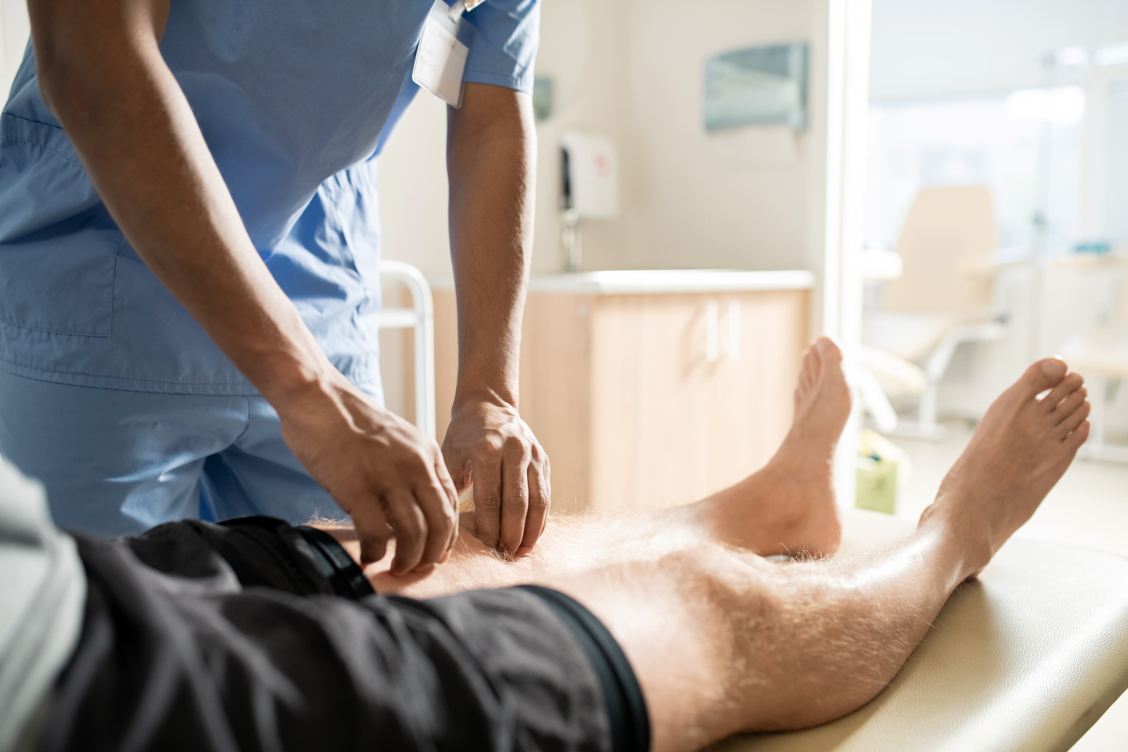 Healthcare worker in scrubs performing a leg examination on a patient lying on a clinical table.