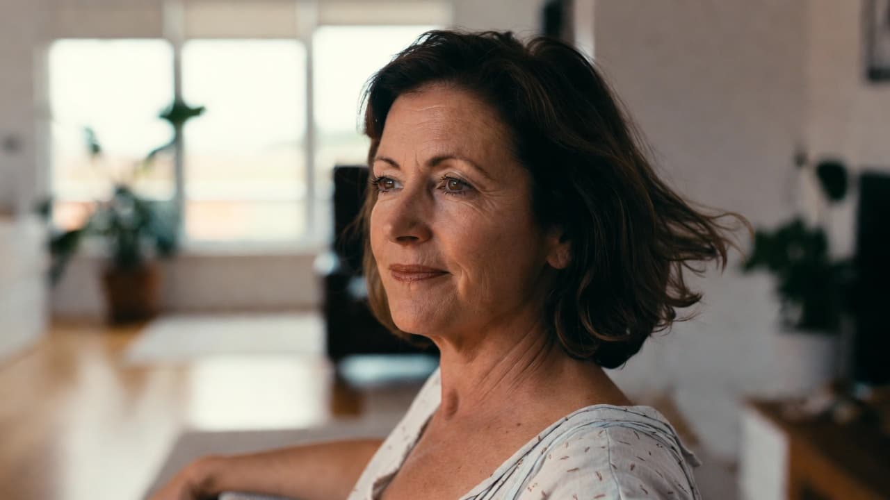 A middle-aged woman with brown hair smiles softly in a warmly lit home interior.